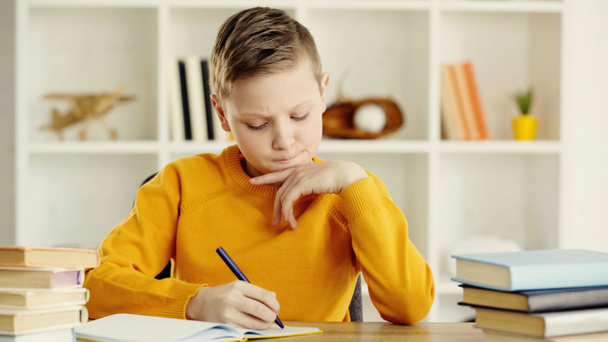 thoughtful preteen schoolboy writing in notebook near books on desk  - Foto, immagini