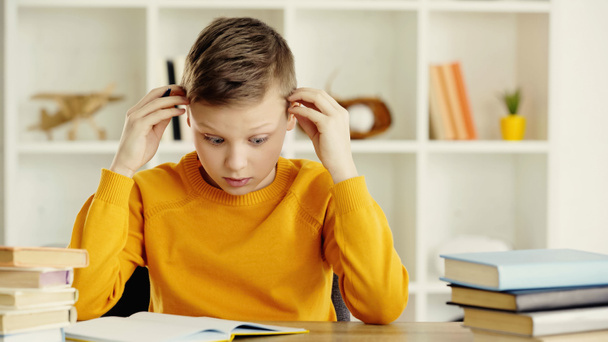 tensed preteen boy looking at notebook while doing homework near books on wooden desk  - Foto, Imagen