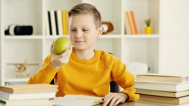 schoolboy smiling while looking at green and tasty apple at home  - Fotó, kép