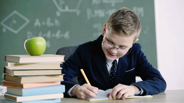 cheerful schoolboy in glasses writing in notebook near apple on books in classroom  - Фото, изображение