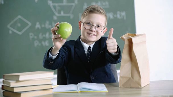 happy schoolboy in glasses holding green apple while showing thumb up near paper bag with lunch in school  - Foto, Imagem