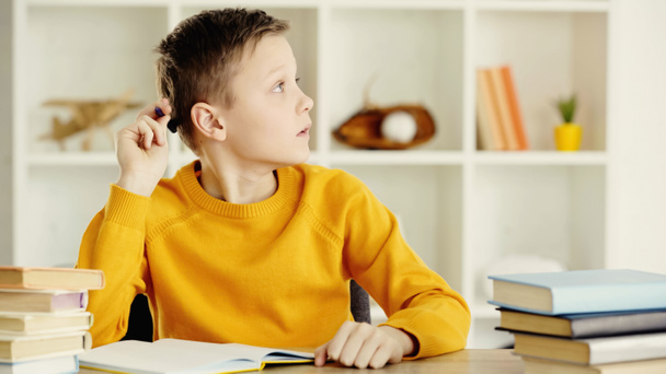 pensive preteen boy thinking while holding pen and doing homework near books on wooden table - Photo, Image