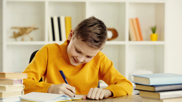 smiling preteen boy writing in notebook while doing homework near books on wooden table at home - Foto, Imagen