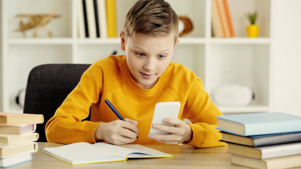 schoolboy looking at smartphone while writing in notebook near books in classroom  - Foto, afbeelding