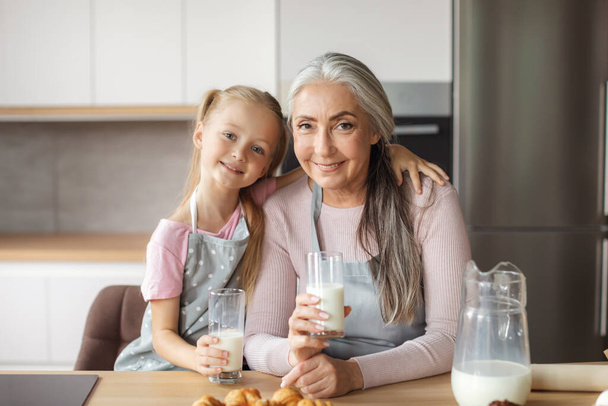 Smiling european old female and little girl in aprons with glasses of milk enjoy croissants in kitchen interior. Healthy breakfast with homemade food, cooking together, baking at home and sweets - Фото, изображение