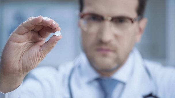 blurred doctor in glasses and white coat looking at camera and holding pill  - Фото, зображення