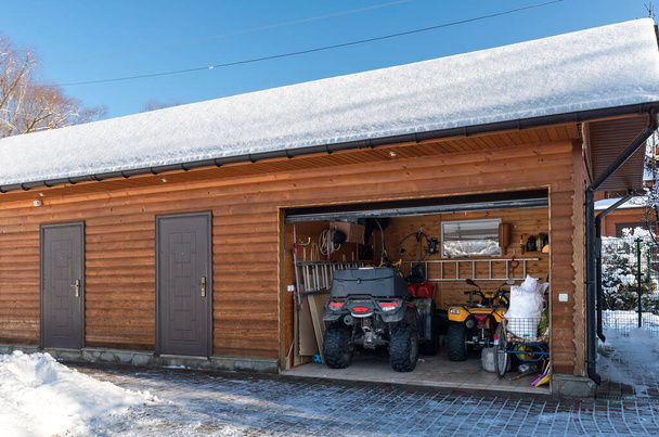 Facade view open door ATV home garage with quad bikes offroad vehicle parked sunny snowy cold winter day. ATV adventure extreme sport. House organized clutter warehouse tools equipment shed storage. - Фото, изображение