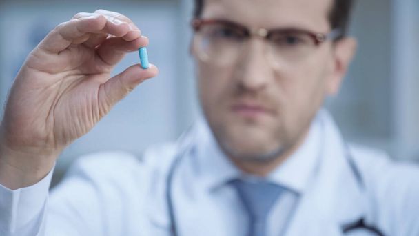 blurred doctor in glasses and white coat looking at camera and holding capsule - Foto, afbeelding