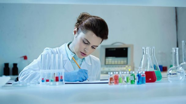 pretty scientist in white coat writing down result near test tubes and flasks with liquid in laboratory - Photo, Image