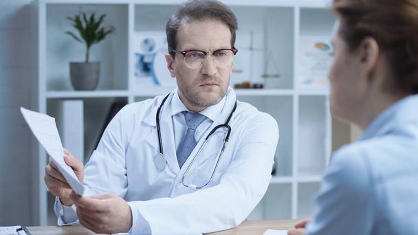 cardiologist in glasses and white coat holding cardiogram and talking to woman in clinic  - Foto, immagini
