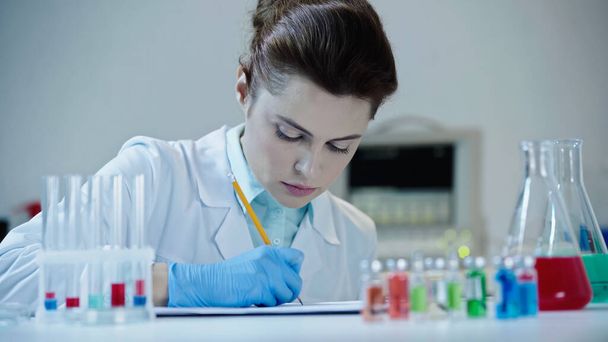 attentive female scientist writing on clipboard result of analysis near test tubes   - Foto, Bild