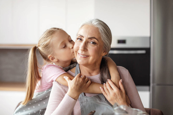 Portrait of smiling european little granddaughter kisses cheek of elderly grandmother in apron in minimalist kitchen interior. Cooking together for family at home, love and relationship, housekeeping - Φωτογραφία, εικόνα
