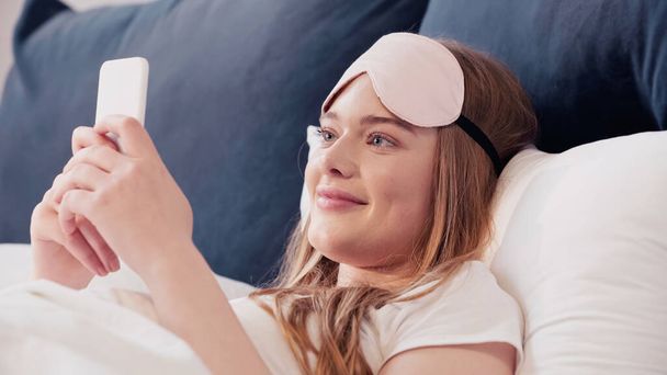 Весела жінка в масці для сну використовує мобільний телефон на ліжку вдома
  - Фото, зображення