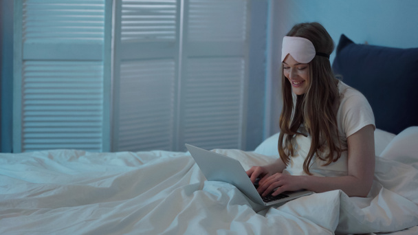 Cheerful woman in sleep mask using laptop on bed at night  - Photo, image