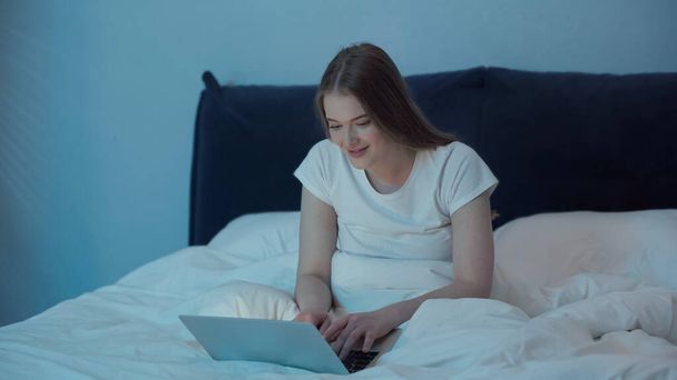 Smiling woman using laptop in bedroom at night  - Fotografie, Obrázek