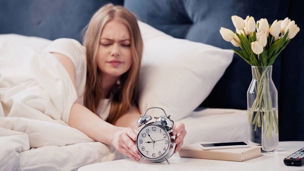 Upset woman touching alarm clock near flowers and smartphone in bedroom  - Foto, afbeelding