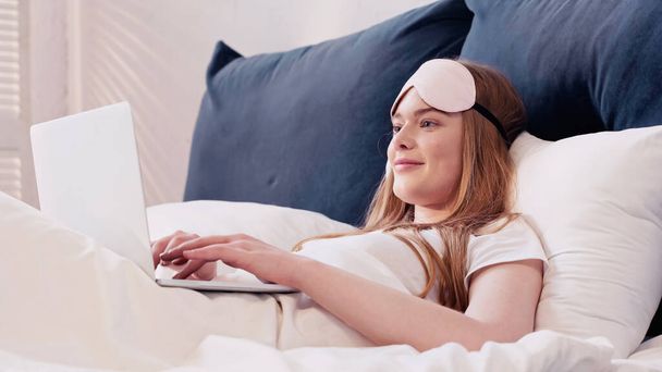 Positive freelancer in sleep mask using laptop while lying on bed in morning  - Φωτογραφία, εικόνα