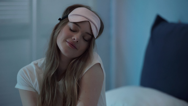 Молода жінка в масці для сну закриває очі, сидячи на ліжку вночі
  - Фото, зображення