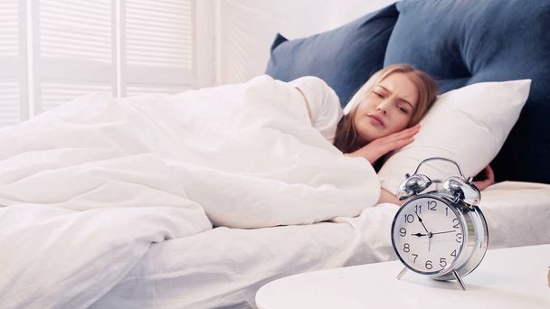 Sad woman looking at alarm clock on bedside table in bedroom - Фото, изображение