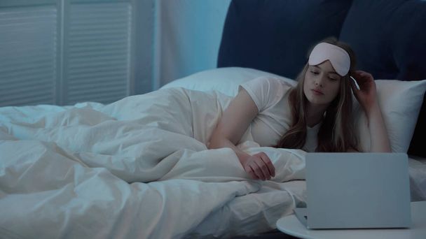 Woman in sleep mask looking at laptop on bedside table in bedroom  - Foto, afbeelding