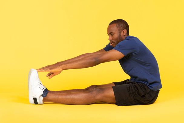 Спортивний афроамериканський чоловік займається стретчінгом, торкаючись до пальців, сидячи над жовтим фоном студії, одягнений в одяг. Фітнес-тренування, концепція здорового навчання. Знімок вигляду збоку
 - Фото, зображення