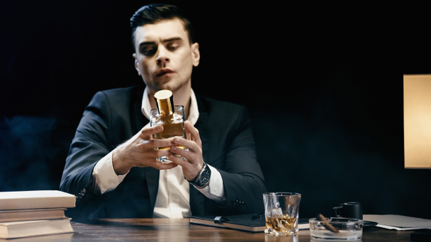 Businessman holding bottle of whiskey near books and cigar on table on black  - Photo, image