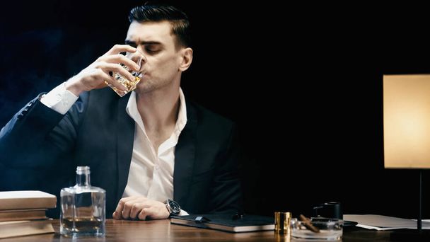 Businessman in suit drinking whiskey near books and ashtray on table isolated on black  - Photo, Image