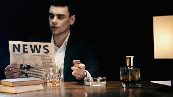 Blurred businessman reading newspaper and holding lighter near ashtray and whiskey on table isolated on black  - 写真・画像
