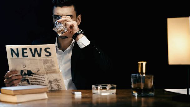 Businessman drinking whiskey and reading newspaper near books and ashtray on table isolated on black  - Foto, Bild
