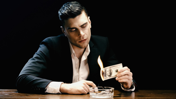 Businessman in suit holding burning dollar banknote near ashtray isolated on black  - Foto, Imagen