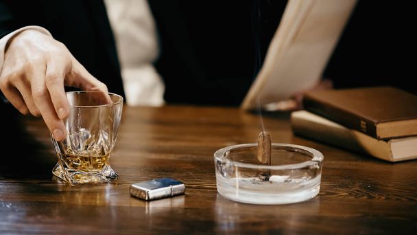 Cropped view of businessman holding glass of whiskey near cigar with smoke in ashtray isolated on black  - Foto, immagini