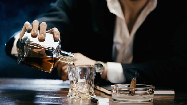 Cropped view of blurred businessman pouring whiskey near cigar in ashtray on black  - Fotó, kép