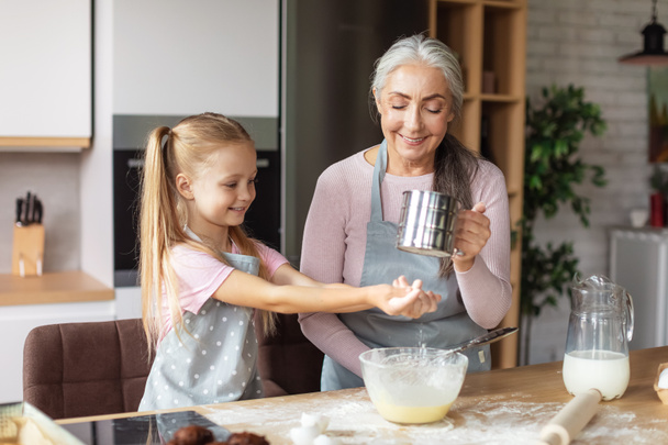 Smiling european little girl and elderly woman in aprons make dough with flour, have fun in kitchen interior. Homemade food, chefs cooking together for family at home, prepare cookies due covid-19 - Foto, Bild