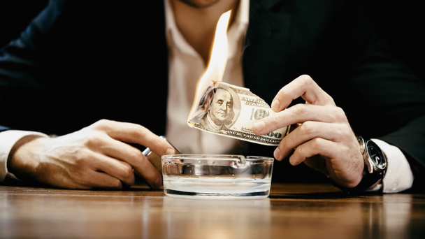 Cropped view of businessman holding burning money and lighter near ashtray isolated on black  - Foto, imagen
