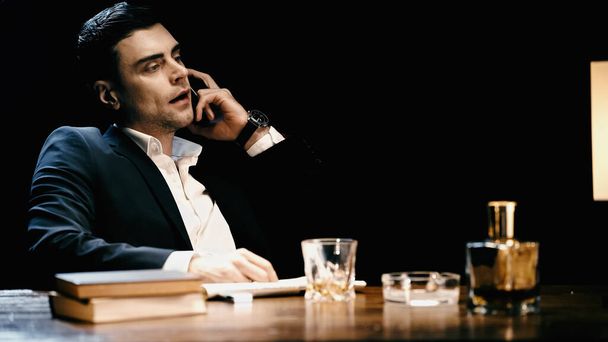 Businessman talking on smartphone near whiskey and books on table isolated on black  - Photo, Image