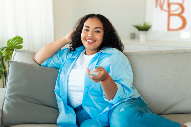 Happy African American Lady Watching Television Pointing Remote Controller At Camera Sitting On Couch At Home. TV Programming, Weekend Leisure And Entertainment Concept - Fotó, kép