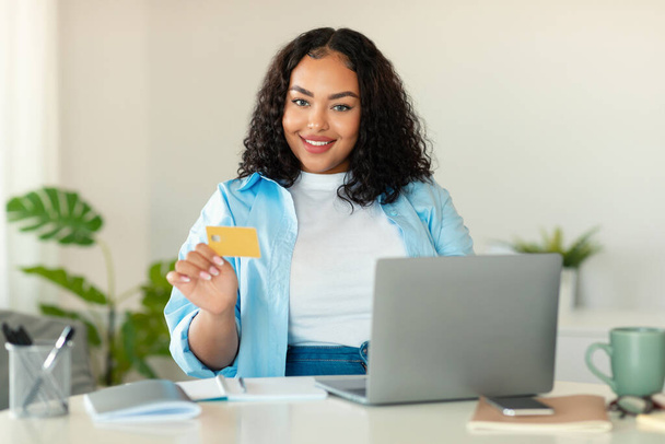 Internet Banking. Happy African American Woman Shopping Online Showing Credit Card Using Laptop Computer Posing Smiling To Camera Sitting In Modern Office. Ecommerce Concept. Selective Focus - 写真・画像