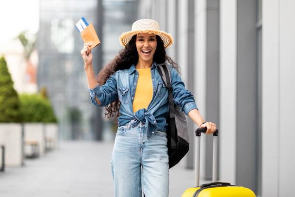 Excited young woman with long curly hair going vacation alone, showing passport with flight tickets, carrying suitcase and backpack, wearing casual outfit and straw hat, city background, copy space - Valokuva, kuva