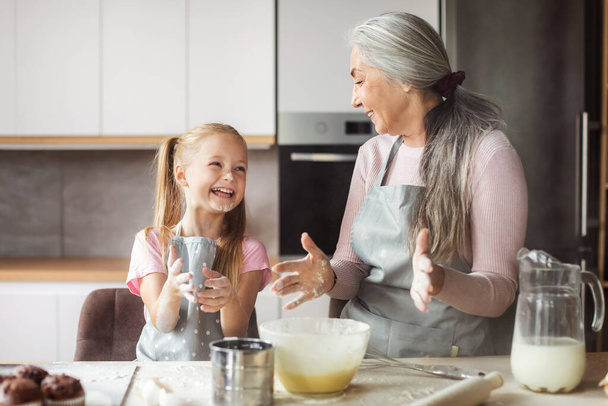 Satisfied european small granddaughter and mature grandmother in aprons make dough with flour, have fun together in kitchen interior. Chefs cooking homemade sweet food at home, love and relationship - Photo, image
