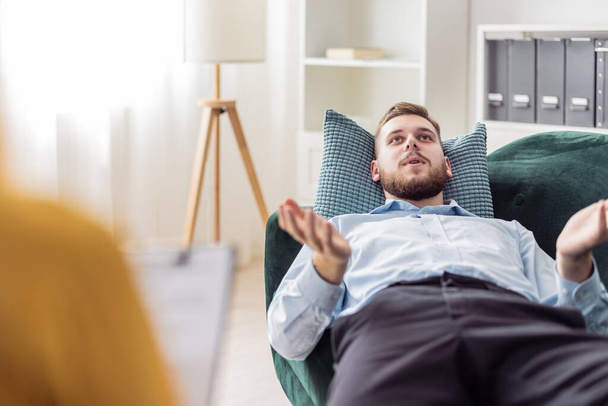Bearded young man talking about strees and problems, psychologist consultation in bright office with sofa, mental health care concept - 写真・画像