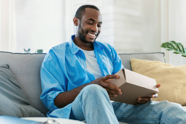 Happy black man unpacking delivery box, sitting on sofa and smiling, male customer opening package, ordering clothes or gadgets on Internet, copy space - Φωτογραφία, εικόνα