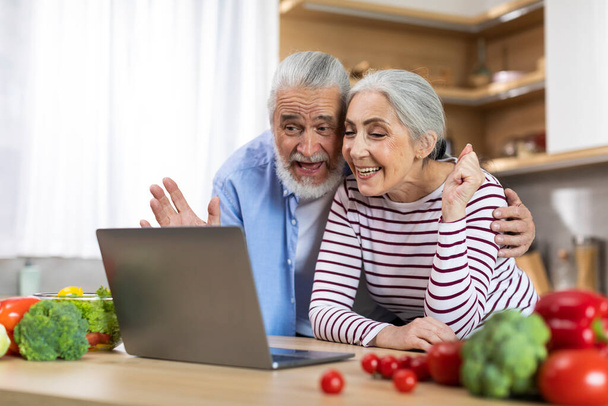 Happy Senior Couple Making Video Call With Laptop While Relaxing In Kitchen, Cheerful Elderly Spouses Waving Hand At Web Camera While Talking To Family, Enjoying Online Communication, Free Space - Foto, immagini