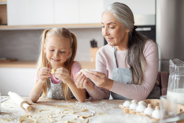 Glad european little girl and old grandmother in aprons make cookies from dough, sculpt dumplings in kitchen interior. Family relationship, lesson of homemade food, prepare with chef together at home - Fotoğraf, Görsel