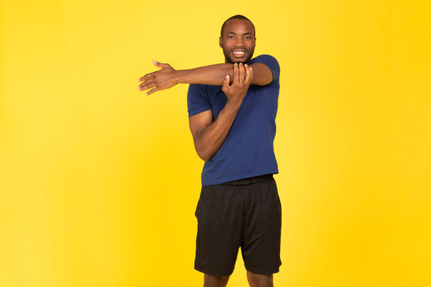 Athletic Black Guy Exercising Stretching Arms Smiling To Camera Having Workout Standing Over Yellow Studio Background, Wearing Sportswear. Fitness Training Concept - Foto, Imagen