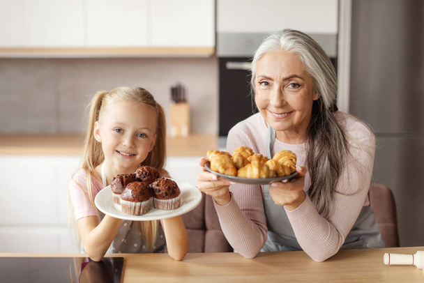 Cheerful pretty little girl and mature grandma in aprons show homemade fresh cookies and croissants in kitchen interior. Cooking baking and prepare food for family at home, household chores together - Foto, imagen