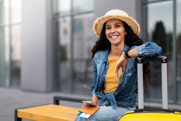 Cheerful young lady tourist in casual outfit and straw hat sitting on bench outside airport, leaning on her big yellow suitcase, holding passport with flight tickets and smartphone, waiting for taxi - Foto, Bild