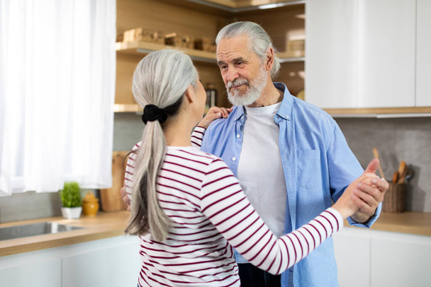 Aged Love. Portrait Of Romantic Elderly Spouses Dancing In Kitchen Interior, Loving Senior Husband And Wife Holding Hands And Looking At Each Other, Enjoying Time At Home Together, Closeup Shot - Φωτογραφία, εικόνα