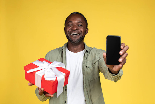 Smiling adult african american man in casual hold gift box and show smartphone with blank screen, isolated on yellow background, studio. Congratulations on holiday, birthday celebration and new app - Фото, изображение