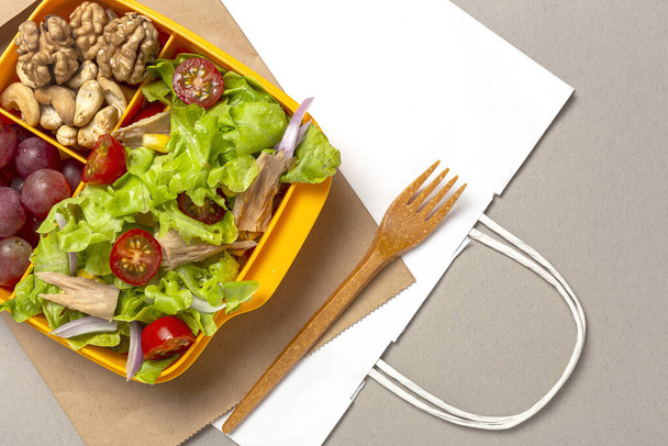 Healthy Food in Plastic container ready to eat with homemade lettuce salad with tomato, onion, corn, tuna, roga grapes, walnuts and cashews in paper bag fro, above. Take away concept - Foto, imagen