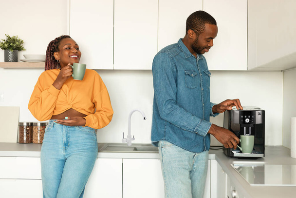 Happy african american couple preparing fresh aromatic coffee with modern machine in kitchen, enjoying hot morning beverage and pleasant talk - 写真・画像